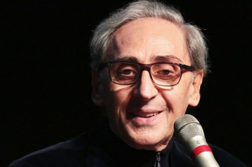 Una foto sorridente di Franco Battiato, su fondo nero, con davanti un microfono